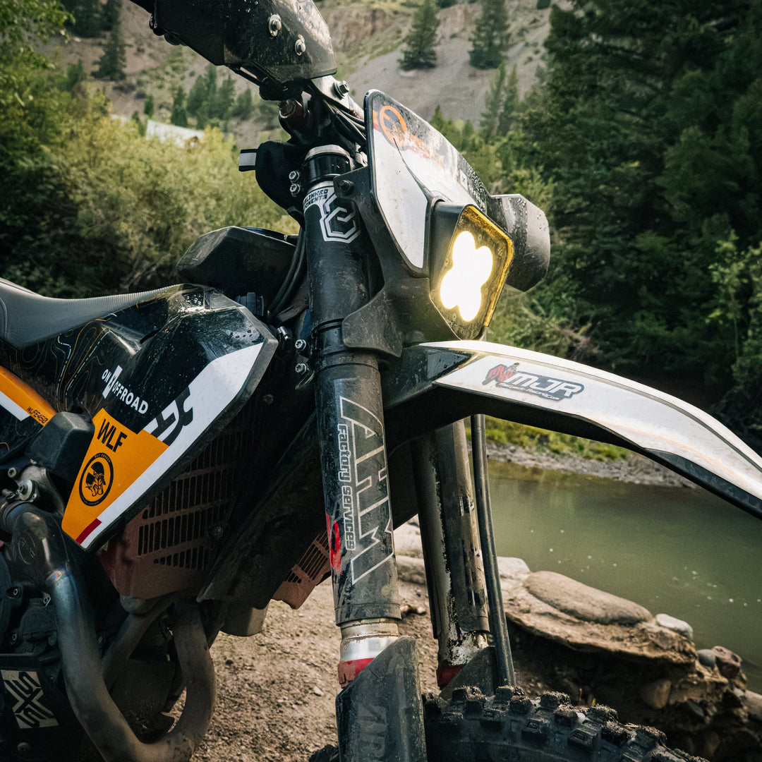 A close-up outdoor shot of a black and white off-road motorcycle with a bright headlight on, parked beside a river with a forested mountain in the background.